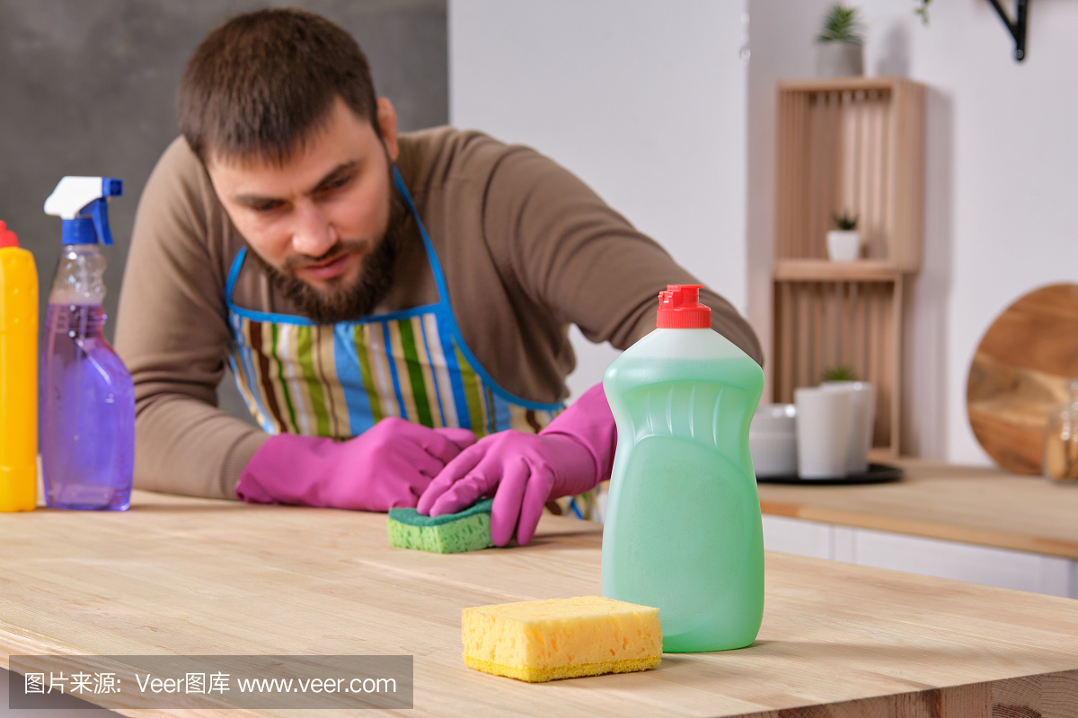 年轻英俊的大胡子男人在厨房里,试图用洗涤剂,刷子,喷雾剂来清理桌子