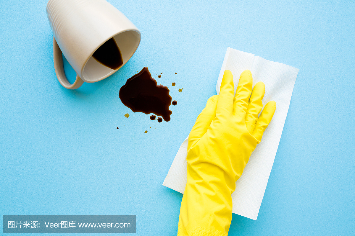 手戴黄色防护橡胶手套清洗新鲜溢出的深色饮料。咖啡渍用白纸巾简单去除。清理。关闭了。浅蓝色的书桌上。前视图。